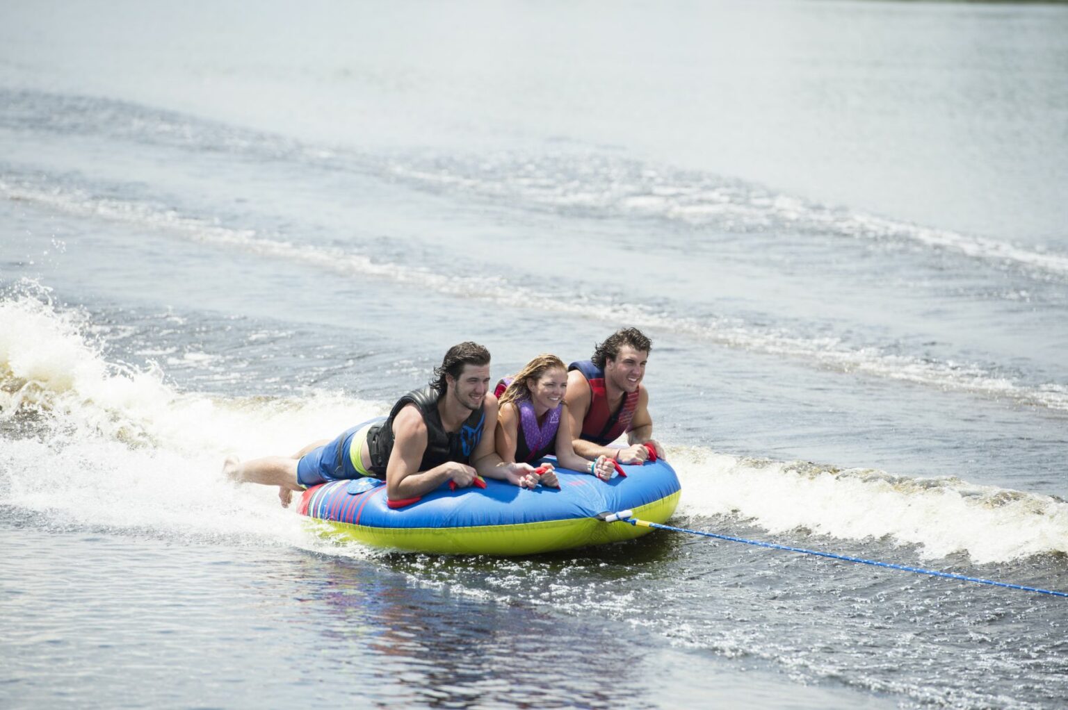 License-free image Three people lie on their stomachs on a colorful inflatable tube being towed by a rope across a body of water. They are smiling and holding onto the handles while riding the waves, creating a fun and dynamic scene.