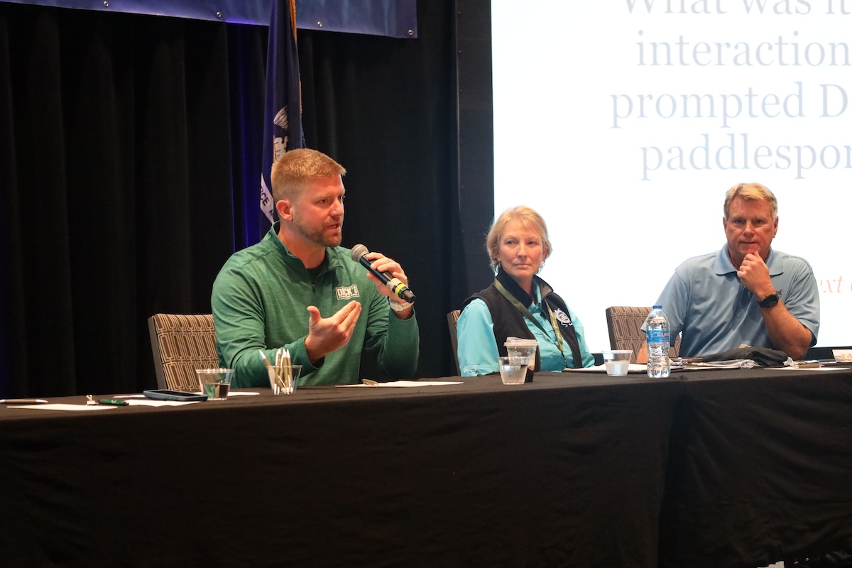 Royalty-free image - Three people sit at a conference panel table. The man on the left speaks into a microphone about the new life jacket law, gesturing emphatically. The woman in the middle and man on the right listen attentively. A presentation screen with text is partially visible behind them.