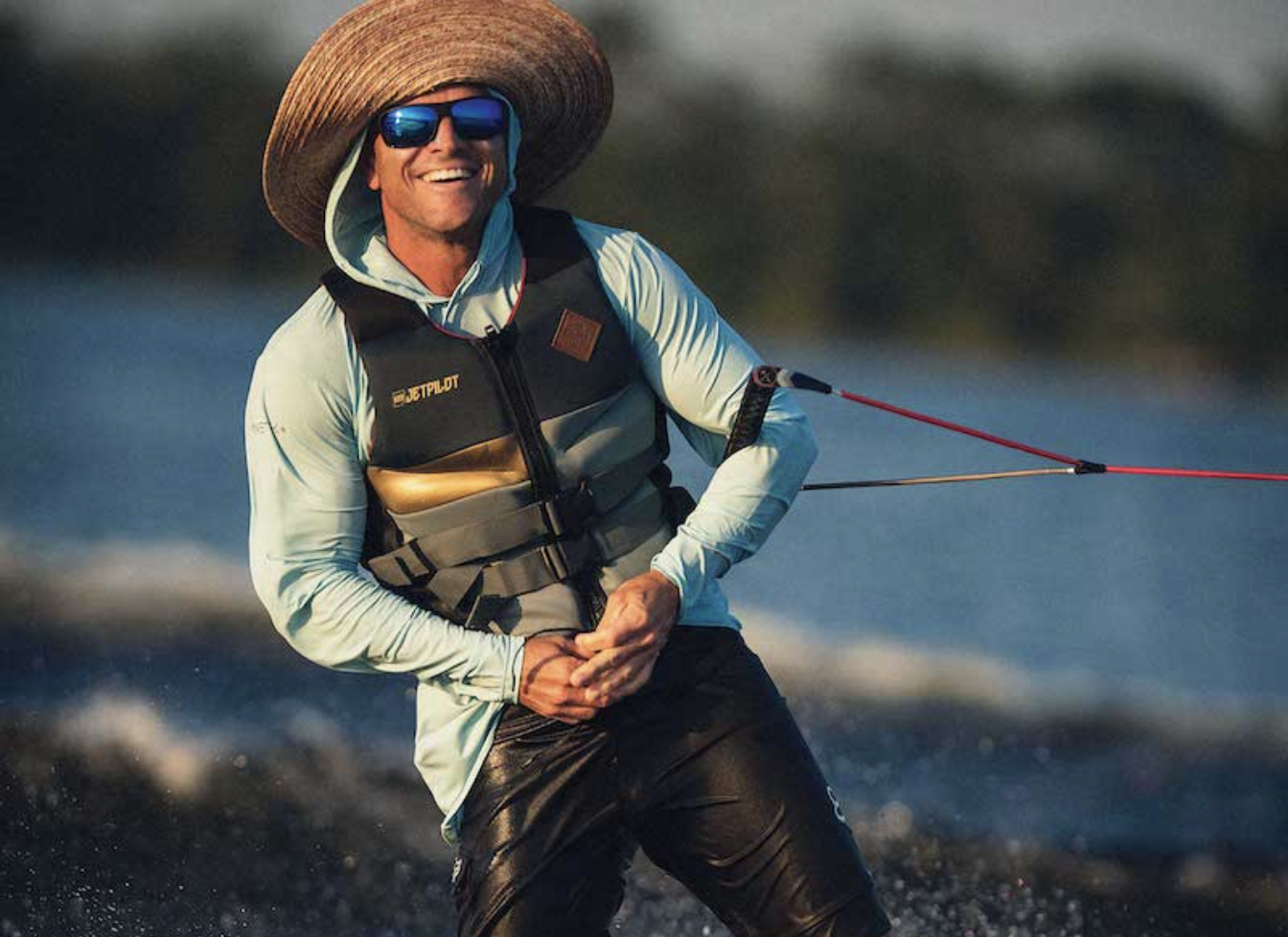 Royalty-free image - A person wearing a large straw hat, sunglasses, and a life jacket is smiling while wakeboarding on a body of water. They are holding onto a tow rope, with blurred water and trees in the background.